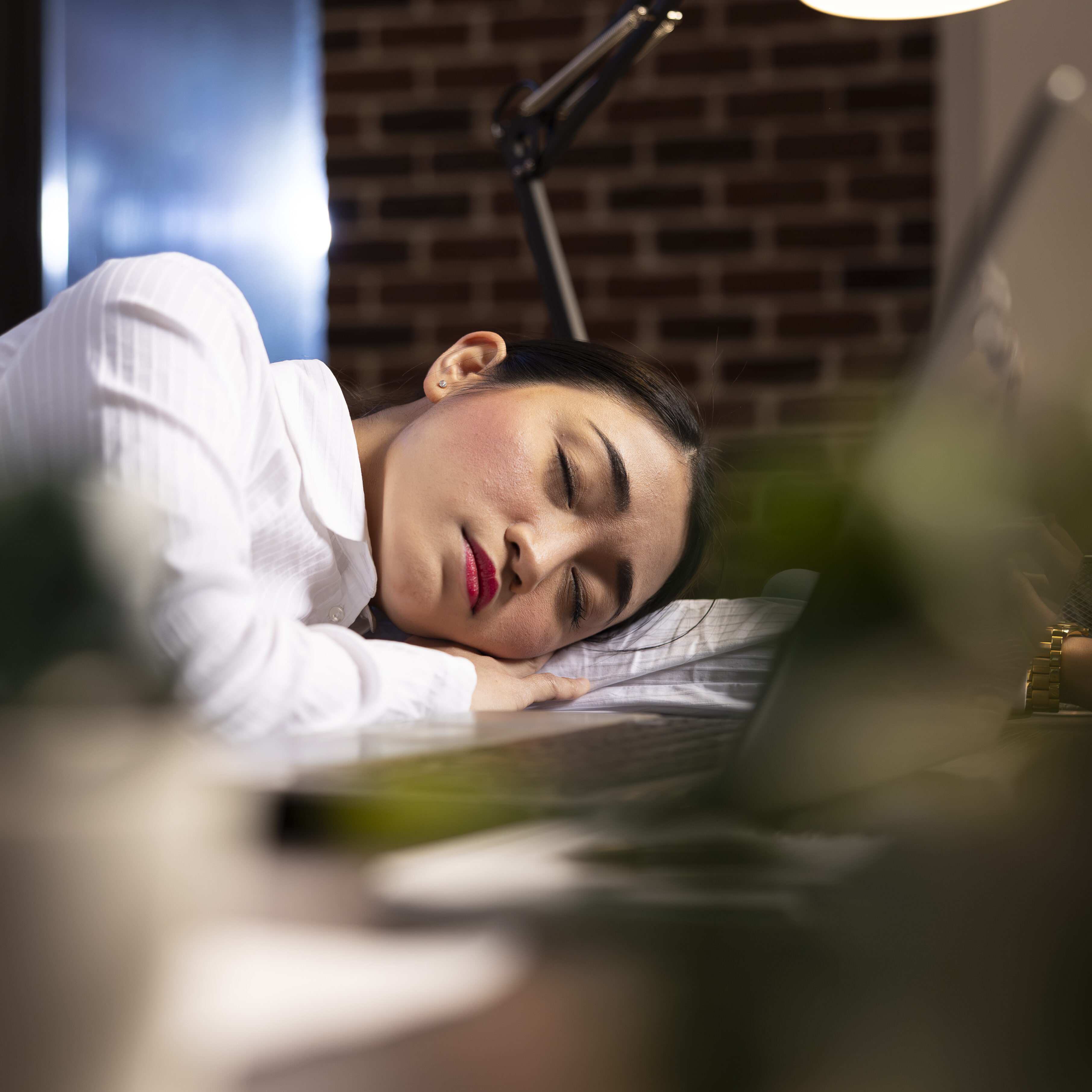 Tired woman manager falls asleep beside her open laptop, taking a powernap during her night shift. Exhausted female employee with eyes closed, resting her head on desk in brick wall office.