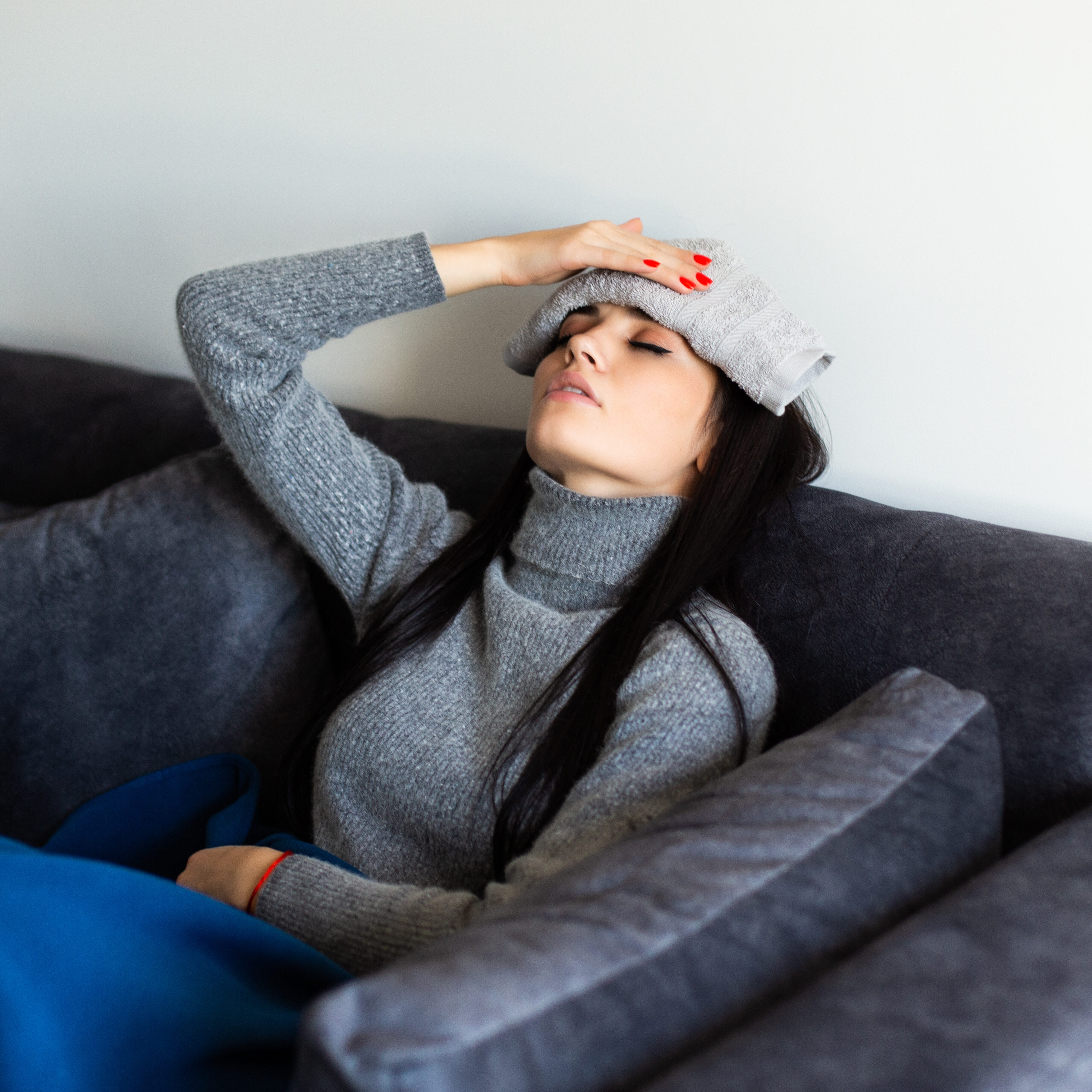 Sick young caucasian woman in grey homewear sitting on bed with laptop working, looking at thermometre after measuring temperature. Illness, freelance concept.