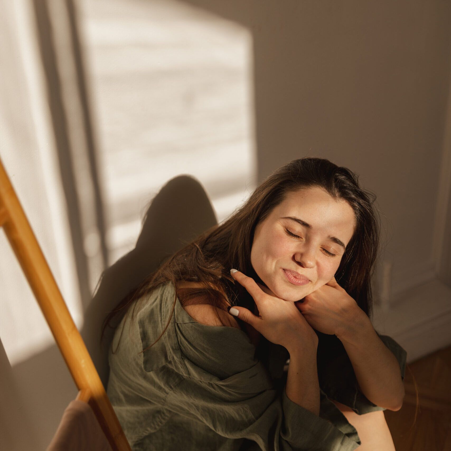 attractive-caucasian-young-girl-smiles-modestly-camera-closing-her-eyes-from-sun-brunette-sits-light-room-against-white-wall-leisure-lifestyle-beauty-concept